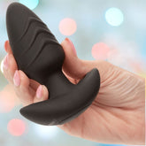 A hand holds the CalExotics Rock Bottom Rotating Probe Vibrating Butt Plug, its black, body-safe silicone ridges highlighted against soft pastel lights—enjoy 10 powerful functions in sleek style.
